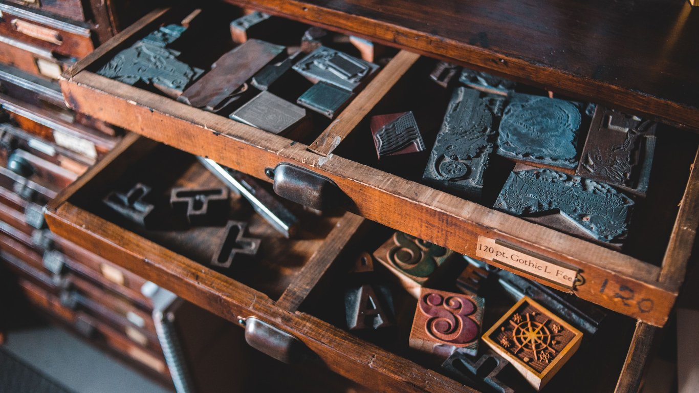 Open wooden drawers filled with vintage metal and wood printing blocks and letterpress tools, photographed in a warm, rustic workshop setting.