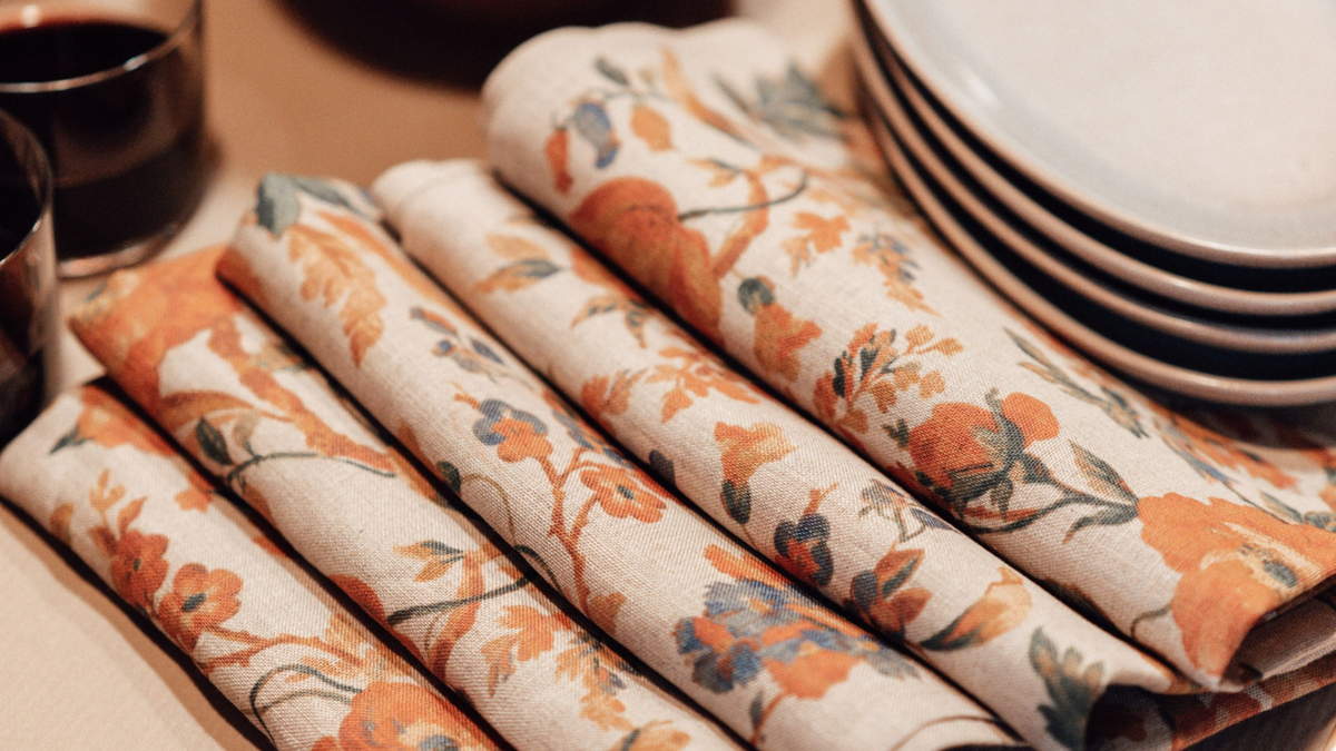 Folded linen napkins with an orange and blue floral pattern on a cream background, arranged on a table next to stacked white plates and a dark glass.