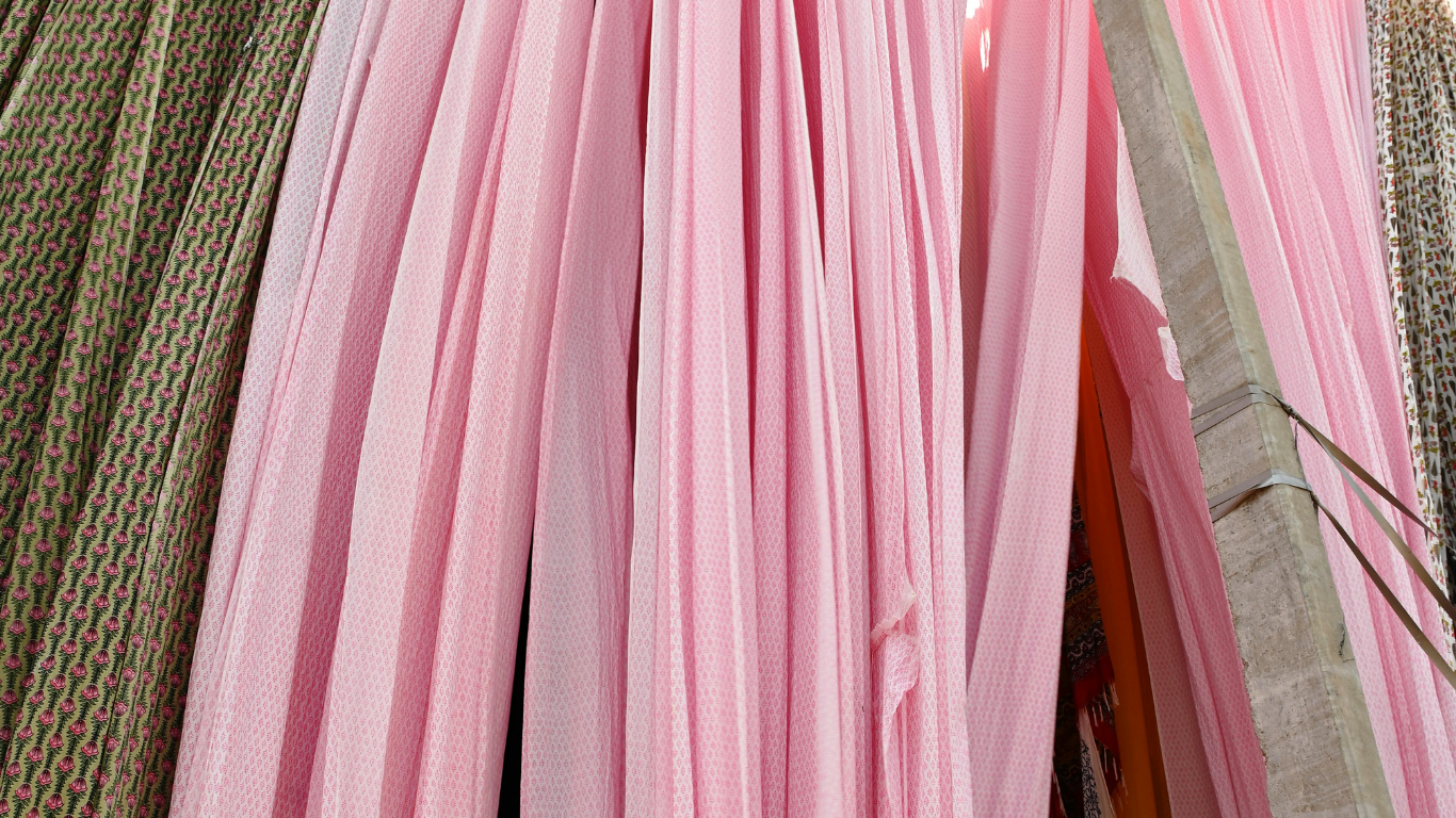 Pink sheer mesh fabric panels hanging vertically, with a textured olive green and red patterned fabric visible on the left side.