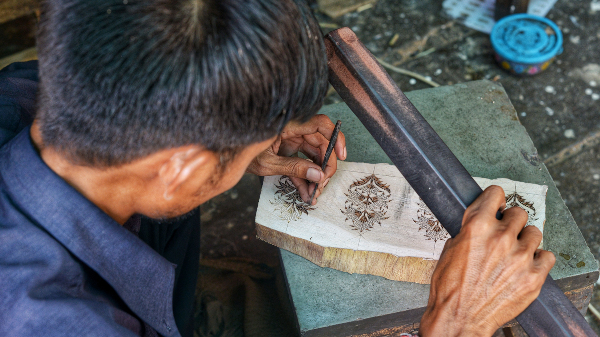 A close-up, over-the-shoulder view of a craftsman carving an ornate floral design into a light-colored wooden block, using a dark metal ruler as a guide on a workbench.